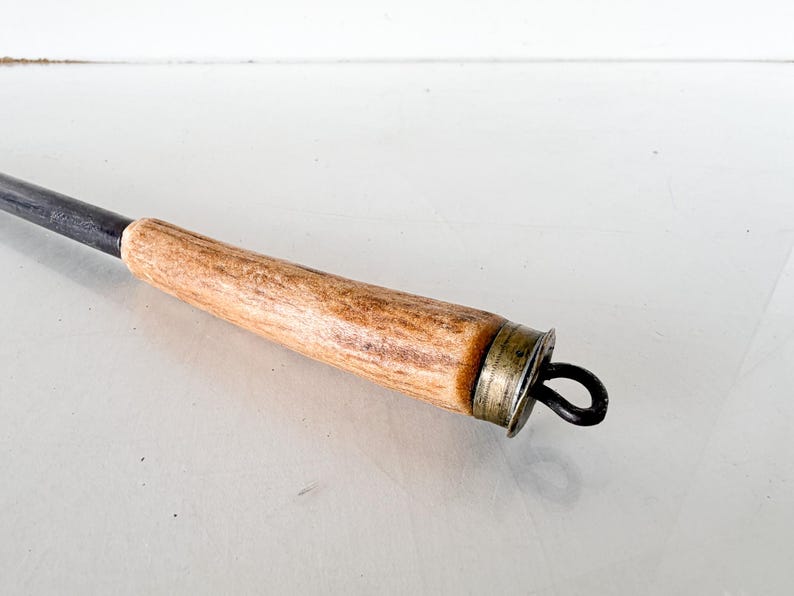 Puede incluir: Primer plano de un atizador de chimenea vintage. El mango es de madera marr&oacute;n claro con un patr&oacute;n texturizado. El extremo met&aacute;lico es oscuro con un lazo para colgar. Un anillo de metal color lat&oacute;n conecta el mango con el atizador.