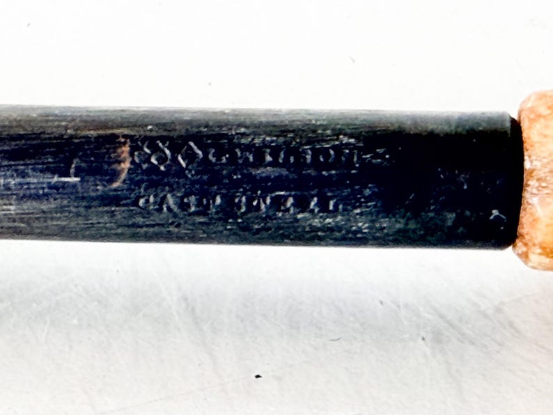 Puede incluir: Primer plano de un mango de madera oscura con texto visible. El mango es negro con un extremo de color claro. El texto parece ser una inscripci&oacute;n, posiblemente una marca del fabricante o informaci&oacute;n de patente.
