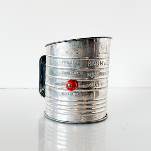 May include: Vintage metal sifter with a black handle and a red knob. The sifter has measurements for five, four, and two cups. The text "BROMWELL" is visible on the side. The sifter is made in the USA.