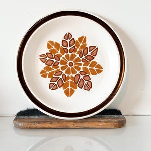 May include: White ceramic plate with a brown rim and a central floral design in shades of brown and orange. The plate is set on a wooden surface, showcasing a vintage aesthetic. This decorative plate is suitable for serving food.