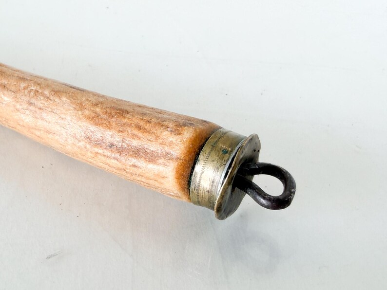 Puede incluir: Primer plano de un mango de madera con una tapa de metal y un lazo de metal oscuro. El mango es de color marr&oacute;n claro con vetas m&aacute;s oscuras. La tapa de metal es plateada y tiene un dise&ntilde;o decorativo.