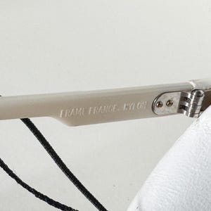 May include: Close-up of a white plastic frame of a pair of glasses with the text "Frame France - Nylon" printed on the side. The frame has a silver metal hinge.