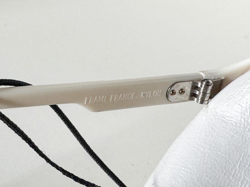 May include: Close-up of a white plastic frame of a pair of glasses with the text "Frame France - Nylon" printed on the side. The frame has a silver metal hinge.