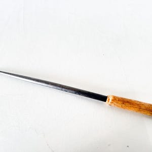 Puede incluir: Una lezna de metal con mango de madera. La lezna tiene una punta larga y afilada y un eje de metal oscuro. El mango es de madera clara y est&aacute; unido al eje de metal con una banda negra.