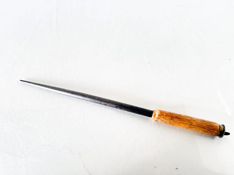 Puede incluir: Una lezna de metal con mango de madera. La lezna tiene una punta larga y afilada y un eje de metal oscuro. El mango es de madera clara y est&aacute; unido al eje de metal con una banda negra.