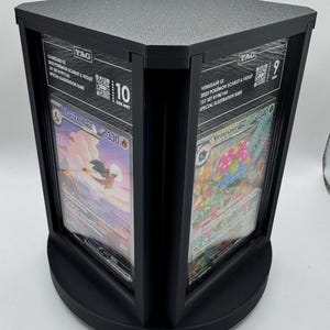 Op de afbeelding: Zwarte displaystandaard voor beoordeelde Pokémon-kaarten. De standaard heeft een vierkante bovenkant en een gebogen basis. Doorzichtige panelen maken het bekijken van de kaarten mogelijk, waaronder Charizard EX en Venusaur EX. De kaarten zijn beoordeeld met 10 en 9.