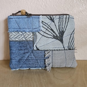 May include: A patchwork zippered pouch featuring denim and patterned fabric in shades of blue, gray, and white. The design includes a botanical print and a wooden bead on the zipper pull. The pouch is rectangular and appears handmade.
