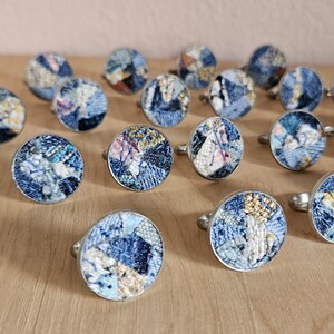 May include: A collection of 15 rings with circular blue, white, and gold fabric inlays. The rings are made of silver metal.