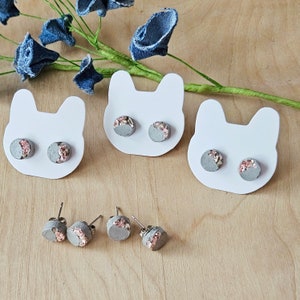 May include: Three pairs of concrete earrings with pink and gold flecks. The earrings are displayed on white cardstock cutouts shaped like cat ears.