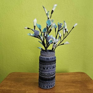 May include: A decorative vase made from denim fabric, holding artificial flowers. The vase is cylindrical, with various denim patterns and stitching. The flowers have white, blue, and teal fabric petals and leaves, with dark stems. The vase sits on a wooden surface.