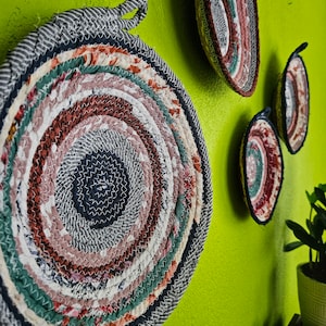 May include: Three round woven wall baskets with a circular pattern of various colors, including pink, green, and brown. The baskets are hanging on a green wall.