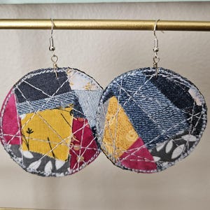 May include: A pair of circular dangle earrings featuring a patchwork design of various fabric scraps in shades of yellow, dark blue, pink, and grey. The fabric pieces are joined with visible white topstitching and hang from silver-toned metal hooks.