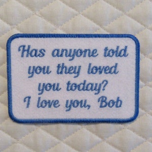 May include: White embroidered patch with blue stitching. The patch has the text "Has anyone told you they loved you today? I love you, Bob".