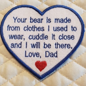 May include: White heart-shaped patch with blue edging, embroidered with the text "Your bear is made from clothes I used to wear, cuddle it close and I will be there. Love, Dad". A small red heart is embroidered in the bottom center.