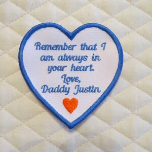 May include: White heart-shaped patch with blue stitching around the edges. The patch has blue embroidered text that reads "Remember that I am always in your heart. Love, Daddy Justin". A small orange heart is embroidered at the bottom of the patch.