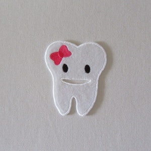 May include: White felt tooth with a pink bow, black eyes, and a stitched smile.