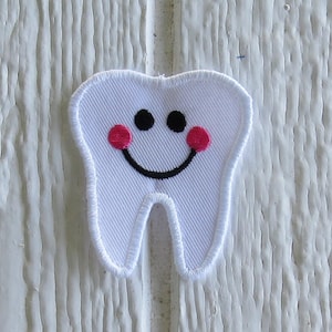 May include: A white tooth-shaped embroidered patch featuring a smiling face with two black circular eyes, a curved black stitched mouth, and two bright pink circular cheeks. The patch has a white stitched border and a ribbed fabric texture.