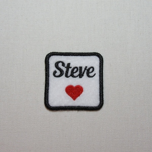 May include: White embroidered patch with black border and black text that says "Steve" with a red heart below the text.