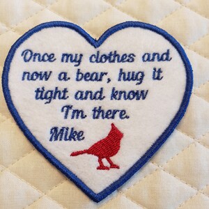 May include: White heart-shaped patch with blue edging. The patch has red embroidered text that reads "Once my clothes and now a bear, hug it tight and know I'm there. Mike". A red cardinal bird is embroidered in the bottom center of the patch.