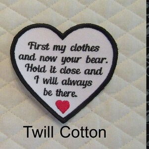 May include: White heart-shaped patch with black stitching and the text "First my clothes and now your bear. Hold it close and I will always be there." A small red heart is stitched in the center of the patch. The patch is on a white quilted fabric background. Twill Cotton is written below the patch.