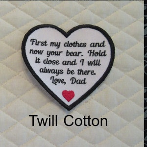 May include: White heart-shaped patch with black embroidered text that reads "First my clothes and now your bear. Hold it close and I will always be there. Love, Dad". A small red heart is embroidered in the bottom center of the patch. The patch is on a white quilted fabric background. The text "Twill Cotton" is printed below the patch.