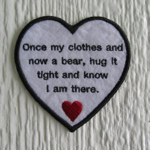 May include: A heart-shaped patch with a white background and a black border. The patch features the embroidered text "Once my clothes and now a bear, hug it tight and know I am there." A small red heart is at the bottom.