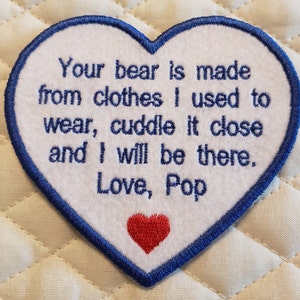 May include: White heart-shaped patch with blue edging. The patch has embroidered text that reads: "Your bear is made from clothes I used to wear, cuddle it close and I will be there. Love, Pop". A small red heart is embroidered in the lower center of the patch.