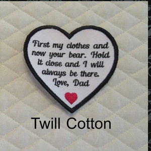 May include: White heart-shaped patch with black embroidered text that reads "First my clothes and now your bear. Hold it close and I will always be there. Love, Dad". A small red heart is embroidered in the bottom center of the patch. The patch is on a white quilted fabric background. The text "Twill Cotton" is printed below the patch.