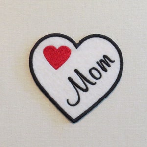 May include: White felt heart-shaped patch with black stitching around the edges. The patch has a red felt heart in the upper left corner and the word "Mom" written in black cursive script.
