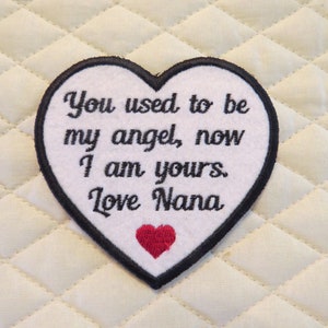 May include: White heart-shaped embroidered patch with black stitching and the text "You used to be my angel, now I am yours. Love Nana" and a small red heart.