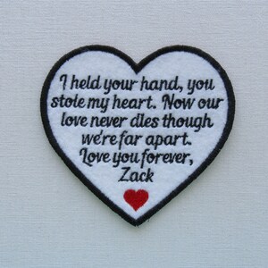 May include: White heart-shaped patch with black stitching and a red heart in the bottom corner. The patch has the text "I held your hand, you stole my heart. Now our love never dies though were far apart. Love you forever, Zack" embroidered on it.