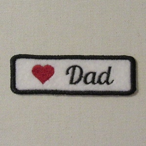 May include: White embroidered patch with a black border. The patch features a red heart and the word "Dad".