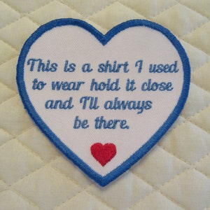 May include: White heart-shaped patch with blue stitching around the edges. The patch has blue embroidered text that reads "This is a shirt I used to wear hold it close and I'll always be there." A small red heart is embroidered at the bottom of the patch.