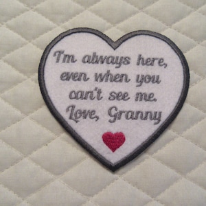 May include: White heart-shaped embroidered patch with gray stitching and the text "I'm always here, even when you can't see me. Love, Granny". A small red heart is embroidered in the bottom center of the patch.