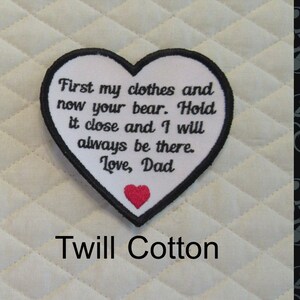 May include: White heart-shaped patch with black embroidered text that reads "First my clothes and now your bear. Hold it close and I will always be there. Love, Dad". A small red heart is embroidered in the bottom center of the patch. The patch is on a white quilted fabric background. The text "Twill Cotton" is printed below the patch.