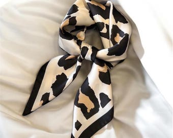 May include: A white silk scarf with a black and brown leopard print design. The scarf is tied in a knot and the ends are hanging down.