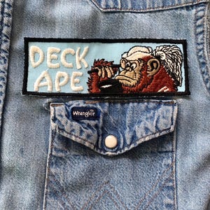 May include: Close-up of a light blue denim shirt with a patch that reads "DECK APE" above a pocket. The patch features a detailed illustration of a monkey. The shirt has a Wrangler label on the pocket.