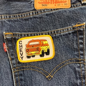 May include: Close-up of dark blue denim jeans with a back pocket featuring a "CHEVY" patch with an orange and yellow truck design. The Levi Strauss & Co. label is visible, along with the "505" model number and waist size 32 and length 30.