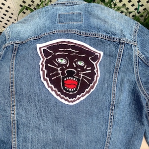 May include: A blue denim jacket with a large patch on the back. The patch features a black panther head with green eyes and a red open mouth. The jacket has a classic design with a collar, button closures, and pockets.