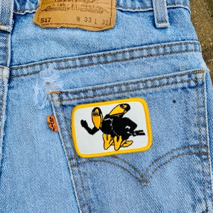 May include: Light blue denim jeans with a distressed pocket and a patch featuring a cartoon bee. The jeans have a classic five-pocket design, a Levi Strauss & Co. label, and a size tag indicating a 33-inch waist and 31-inch inseam.