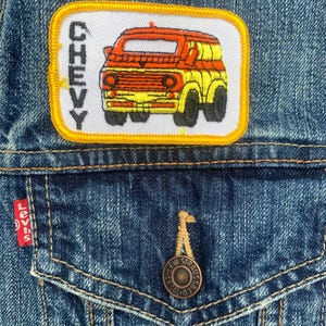 May include: Close-up of a blue denim jacket with a "CHEVY" patch featuring an orange and yellow van design. The jacket has a Levi's tag and a button closure. The patch is rectangular with a yellow border.