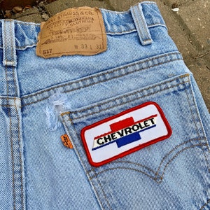 May include: Light blue denim jeans with a distressed pocket and a Chevrolet patch. The patch is white with red and blue stripes and the word "CHEVROLET". The Levi Strauss & Co. tag is visible. Waist size is 33 inches, inseam 31 inches.