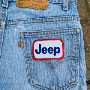 May include: Light blue denim jeans with a distressed pocket. The jeans feature a "Jeep" patch on the back pocket and a Levi Strauss & Co. label. Waist size is W33, and length is L31.