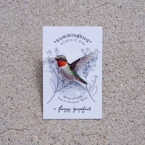 May include: A clear acrylic pin featuring a detailed hummingbird with red, green, and brown coloring. The pin is attached to a white card with the text "hummingbird" and "ACRYLIC PIN". The card also includes the text "designed with love in Houston, Texas" and the brand name "fuzzy grapefruit".
