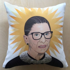 May include: A white pillow with a yellow starburst design and a portrait of Ruth Bader Ginsburg. She is wearing a black robe and a white lace collar.