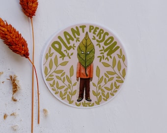 May include: A round sticker with a green leaf covering a person's face. The sticker has a pink background with green leaves and the text "Plant Person" in green.