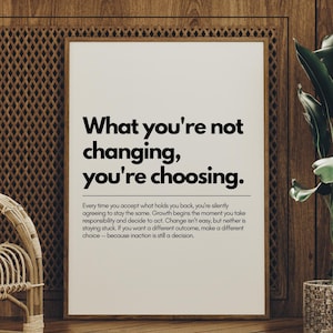 May include: A framed print with the text "What you're not changing, you're choosing." in a black font. The print is displayed against a wooden wall, with a wicker chair and a potted plant in the foreground.