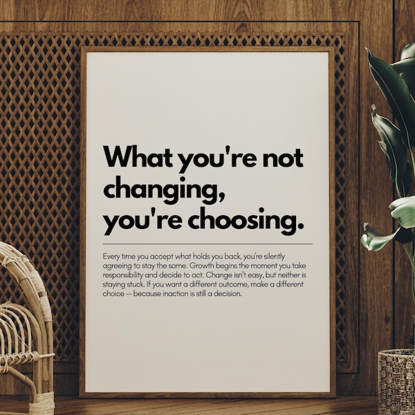 Puede incluir: Un cartel enmarcado con el texto "What you're not changing, you're choosing." en negro. El cartel se muestra contra una pared de madera, con una silla de mimbre y una planta en maceta en primer plano.