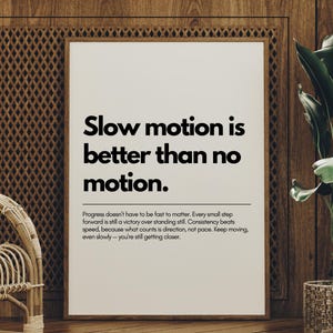 May include: Framed print with the text "Slow motion is better than no motion." The motivational poster is in a white frame, set against a wooden wall. A wicker chair and a potted plant are in the foreground, adding to the room's decor.