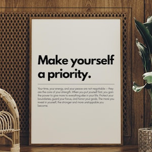 May include: A framed print with the text "Make yourself a priority." in bold black letters. The print is on a white background and is surrounded by a wooden frame. The print is displayed in a room with wooden paneling and a potted plant.